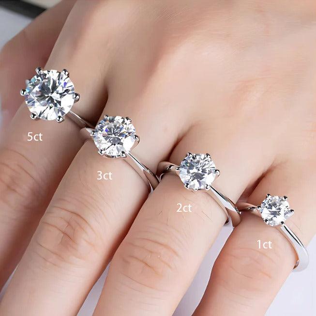 Mão com quatro anéis de prata com diamantes de 1ct, 2ct, 3ct e 5ct em fundo branco.