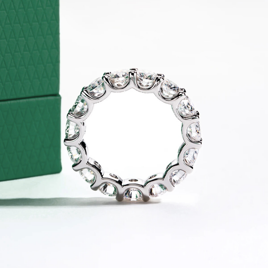 Aliança de prata com pedras de moissanite brilhantes dispostas em círculo, sobre superfície branca ao lado de caixa verde texturizada
