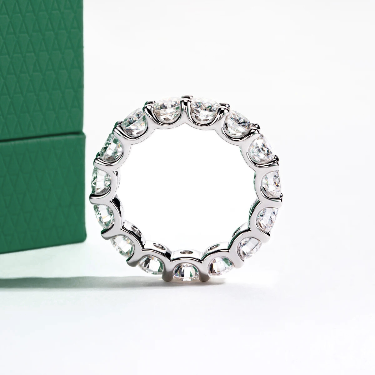 Aliança de prata com pedras de moissanite brilhantes dispostas em círculo, sobre superfície branca ao lado de caixa verde texturizada